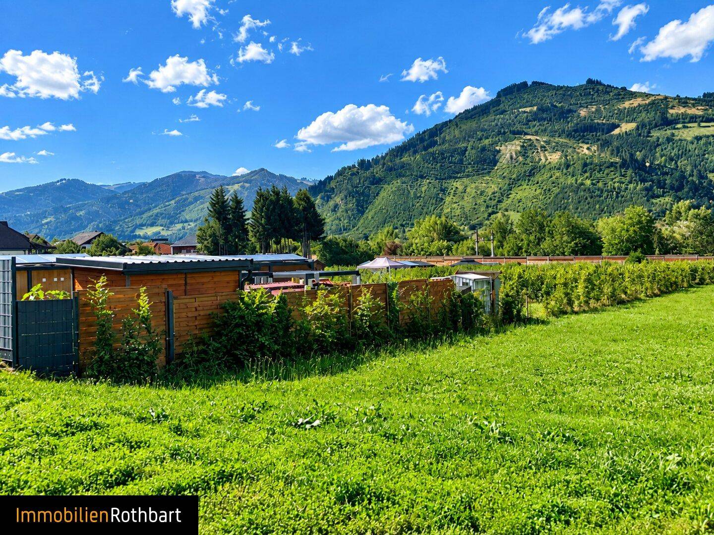 Grüne Rasenfläche mit einem kleinen Holzgebäude und Hecken, umgeben von einer Berglandschaft.