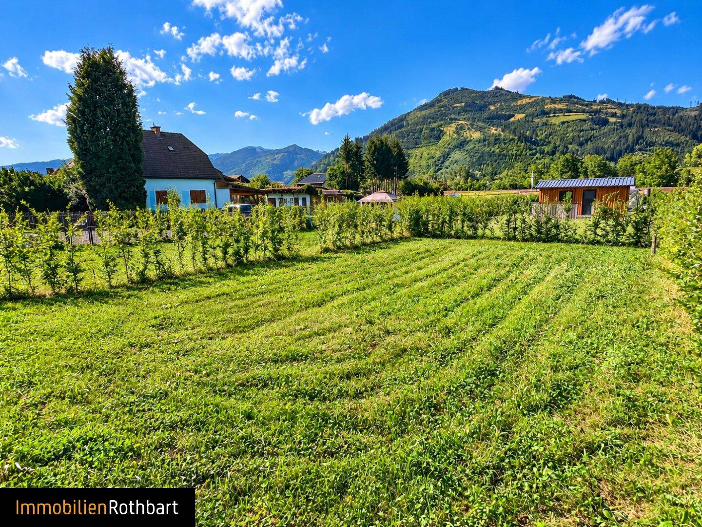 Weitläufige Rasenfläche mit niedrigen Hecken und einem Haus im Hintergrund, umgeben von Bergen.