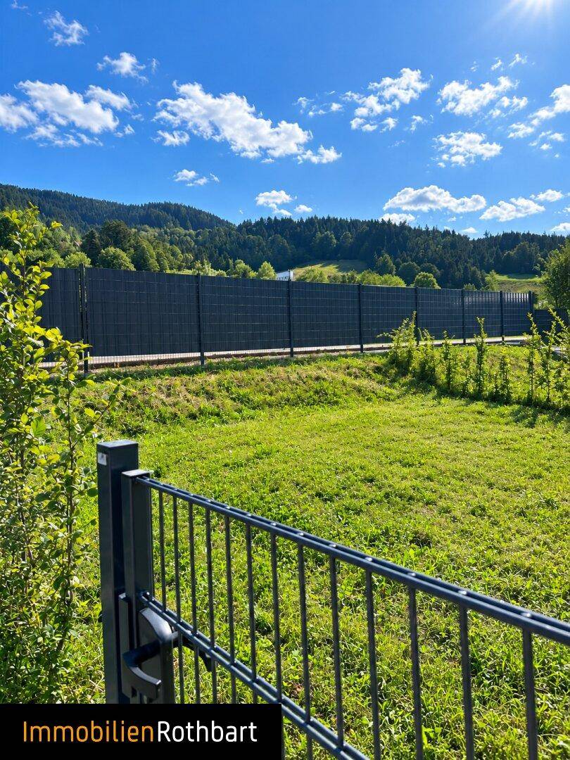 Grüne Wiese mit einem Metallzaun im Vordergrund und einem hohen Sichtschutzzaun im Hintergrund, umgeben von Bergen.