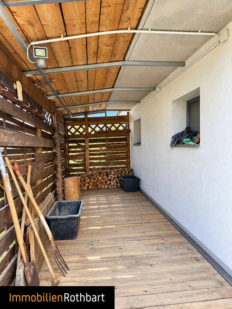 Überdachter Außenbereich mit Holzverkleidung und Lagerplatz für Brennholz und Gartengeräte.