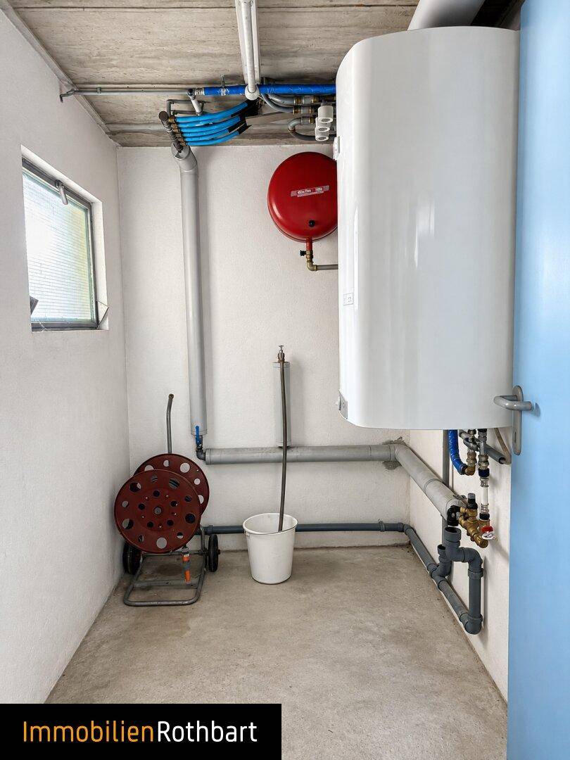 Heizungsraum mit Warmwasserspeicher, sichtbaren Rohren und kleinem Fenster.