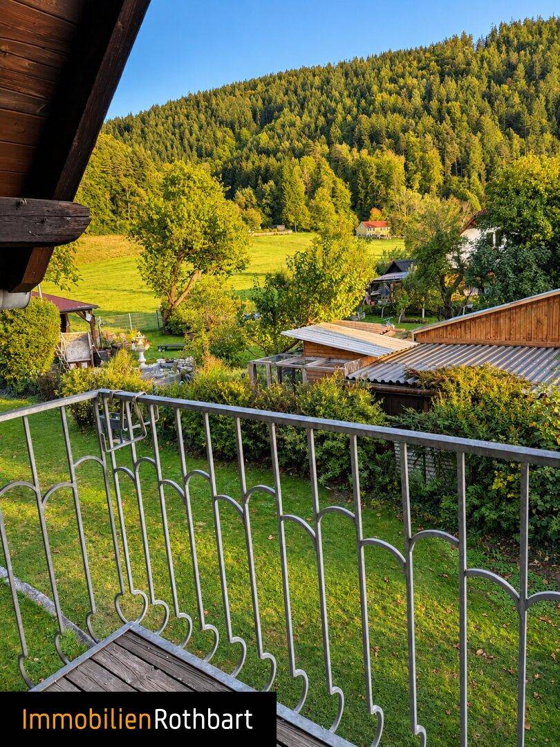 Blick von einem Balkon mit schmiedeeisernem Geländer auf den grünen Garten und die bewaldete Landschaft.