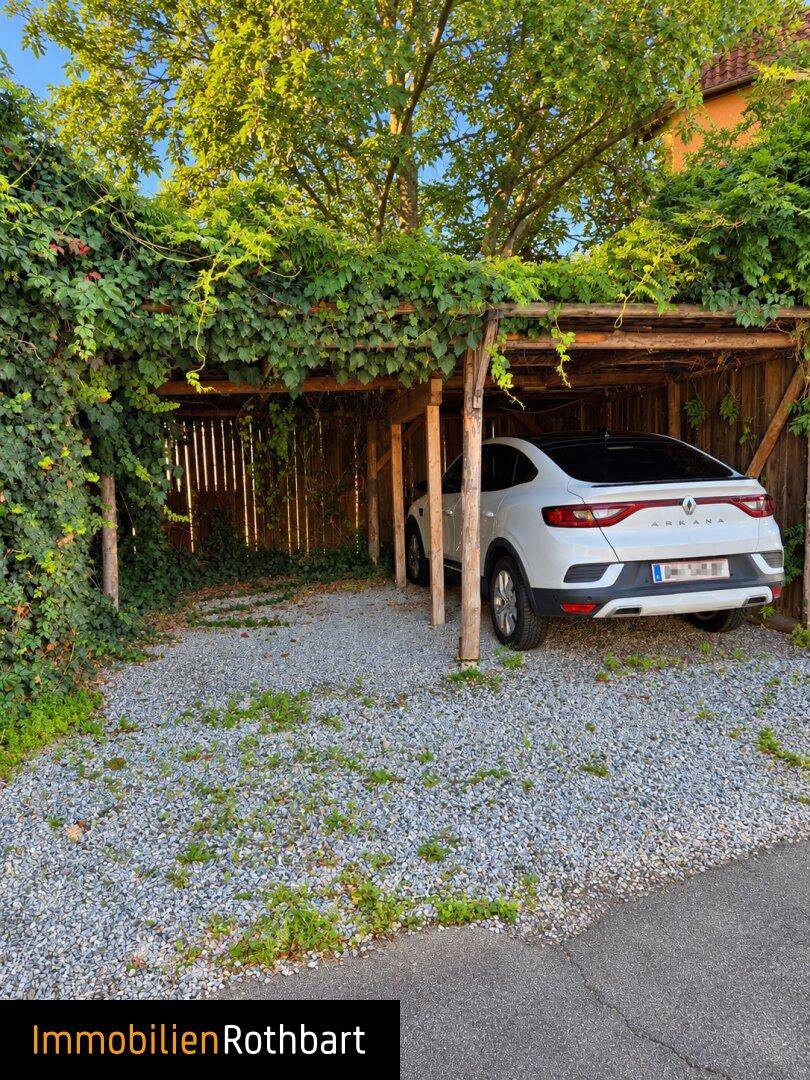 Überdachter Carport mit einem geparkten Auto, umgeben von Rankpflanzen und Schotterboden.