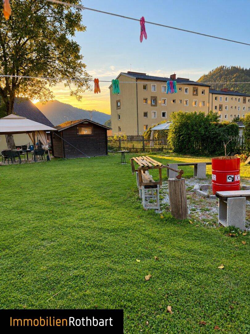 Gartenbereich mit Rasen, Gartenhaus und einer Feuerstelle, im Hintergrund sind weitere Gebäude sichtbar.