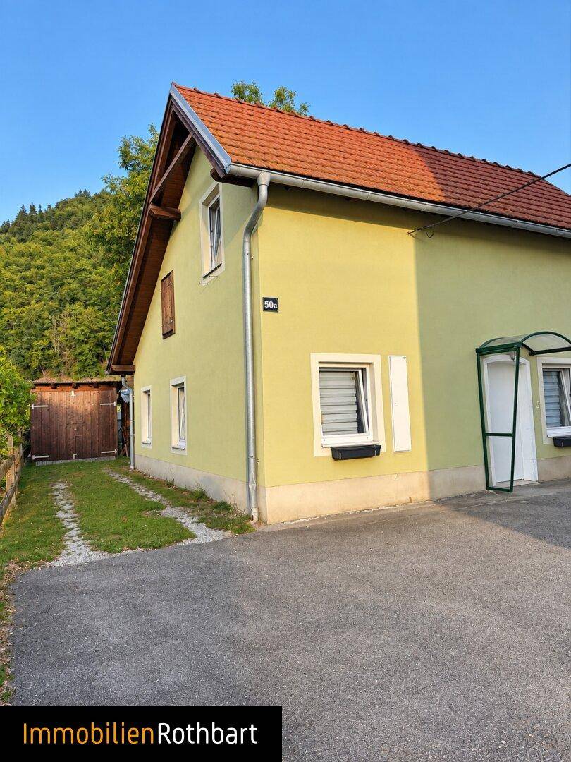 Seitenansicht eines Hauses mit grünlicher Fassade, Satteldach und einem Gartenhaus im Hintergrund.