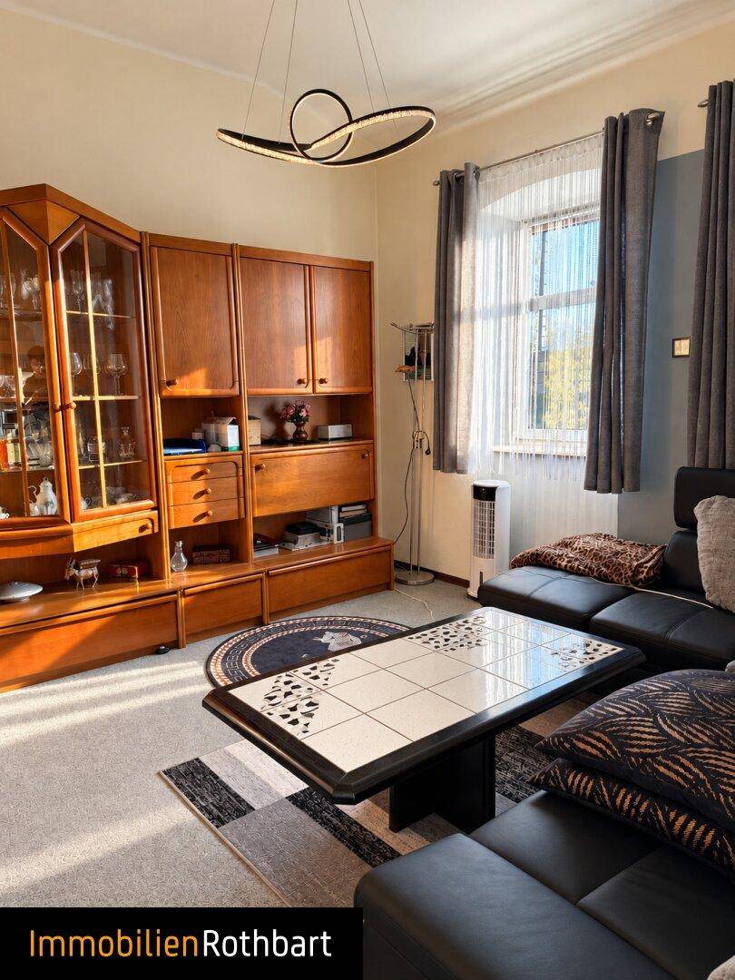 Wohnzimmer mit Teppichboden, dunklem Sofa und einem Holzschrank, beleuchtet durch ein Fenster.