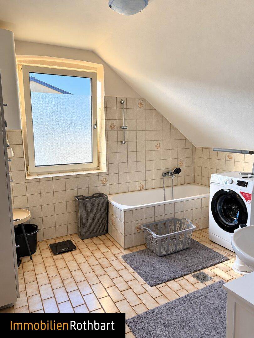 Badezimmer im Dachgeschoss mit Badewanne, Waschmaschine und Fenster, das Tageslicht spendet.