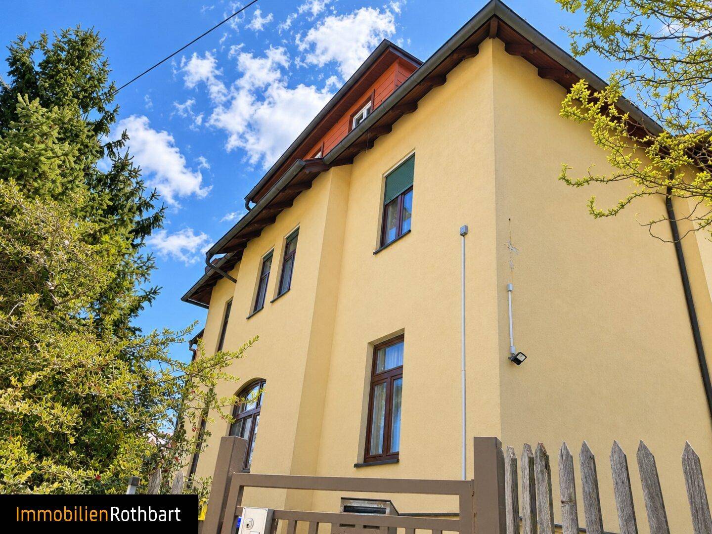 Gelbe Hausfassade mit braunen Fenstern und einem Satteldach unter blauem Himmel.