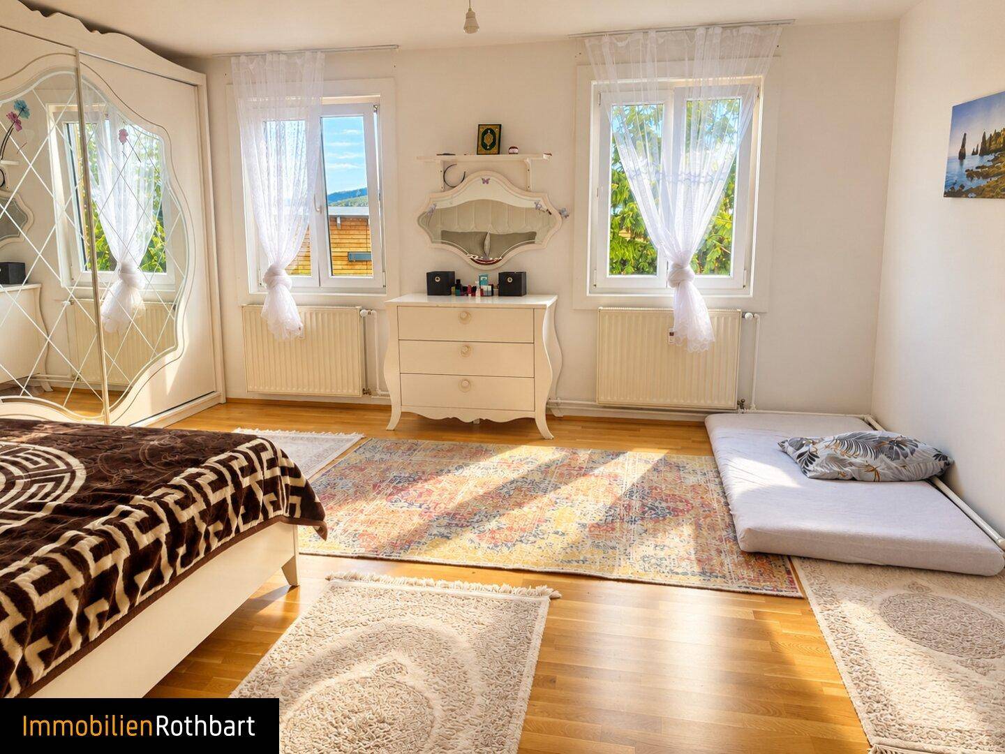 Helles Schlafzimmer mit zwei Fenstern, Holzboden und einem großen Spiegelschrank.