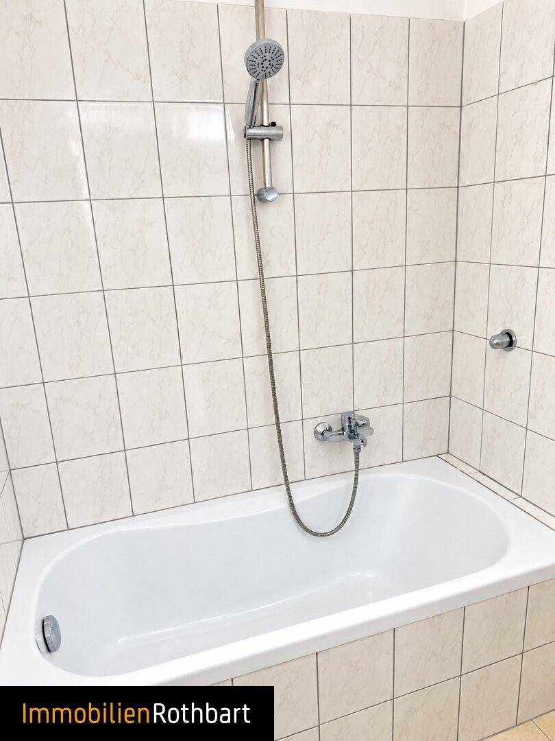 Badezimmer mit Badewanne und Duschvorrichtung, umgeben von hellen Wandfliesen.