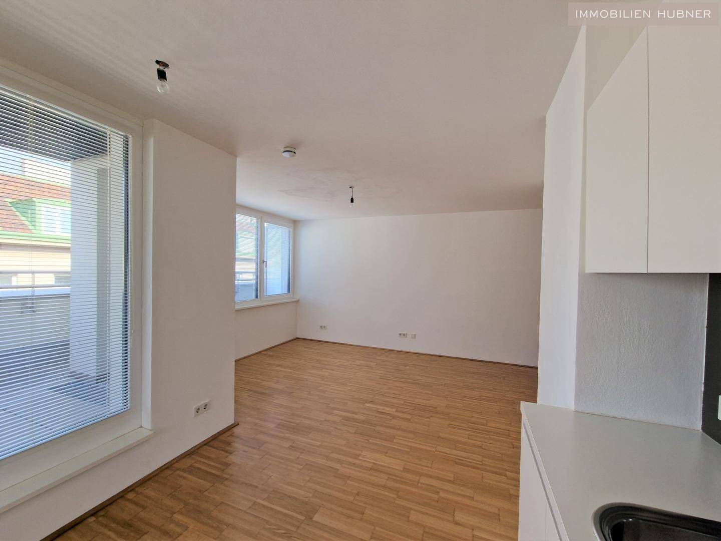Blick von der Küche in den hellen Wohnbereich mit großen Fenstern und Parkettboden.
