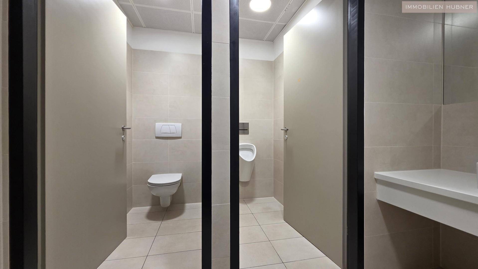 Moderne Toilettenanlage mit mehreren Kabinen, Urinal und einem Waschbeckenbereich mit Spiegel.