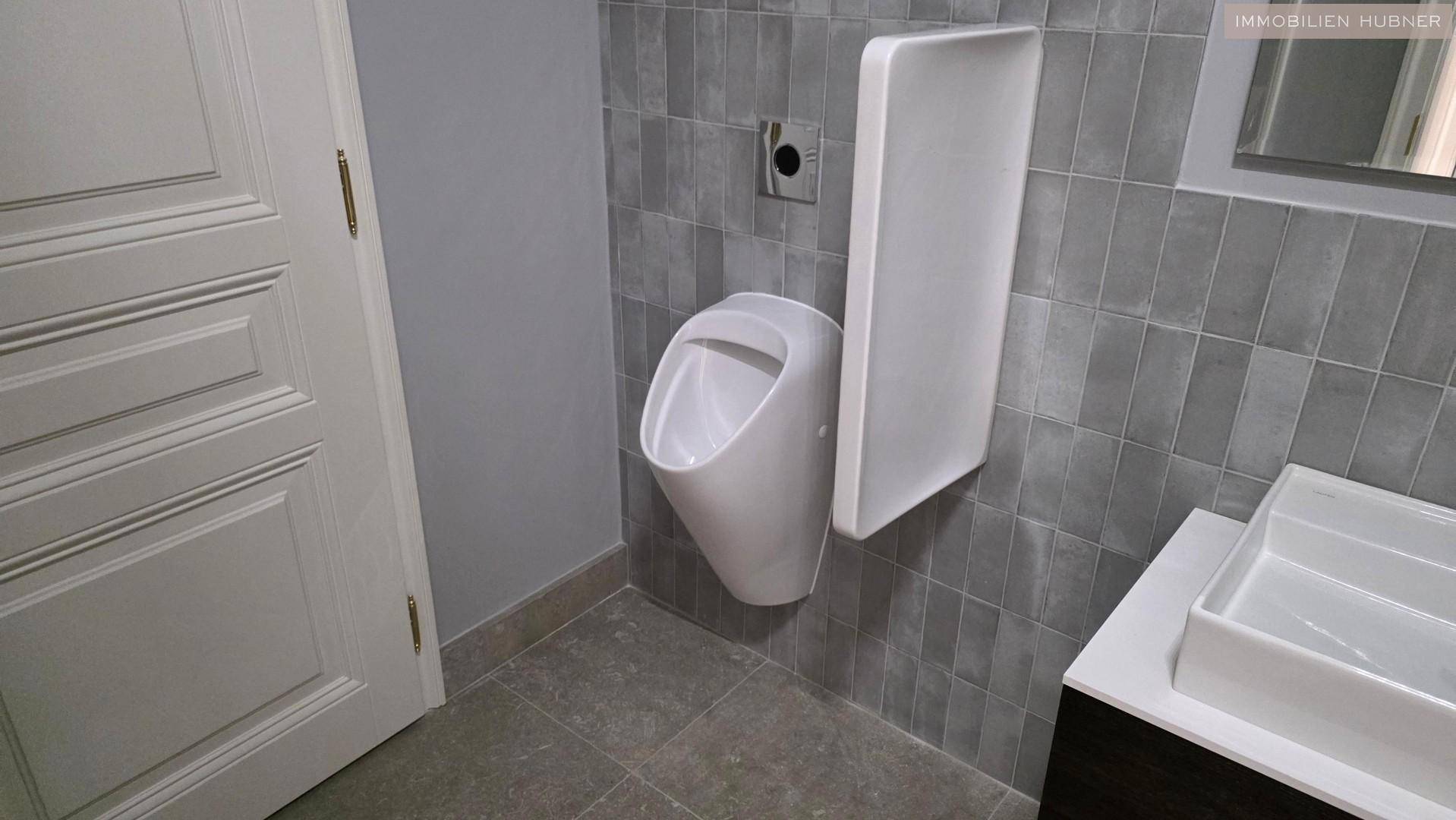 Modernes Badezimmer mit Urinal, Waschbecken und grauen Wandfliesen.