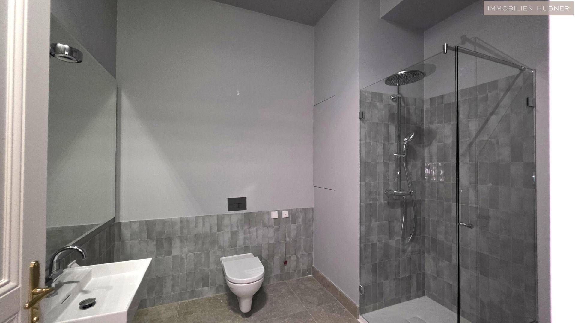 Modernes Badezimmer mit großer Glasdusche, Wand-WC und grauen Fliesen.