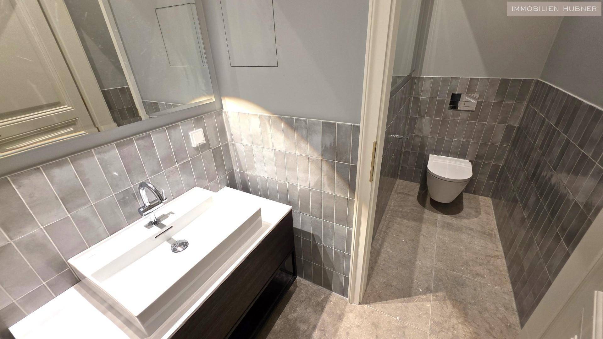 Modernes Badezimmer mit grau gefliesten Wänden und Boden, ausgestattet mit einem stilvollen Waschbecken und einer Wandtoilette.