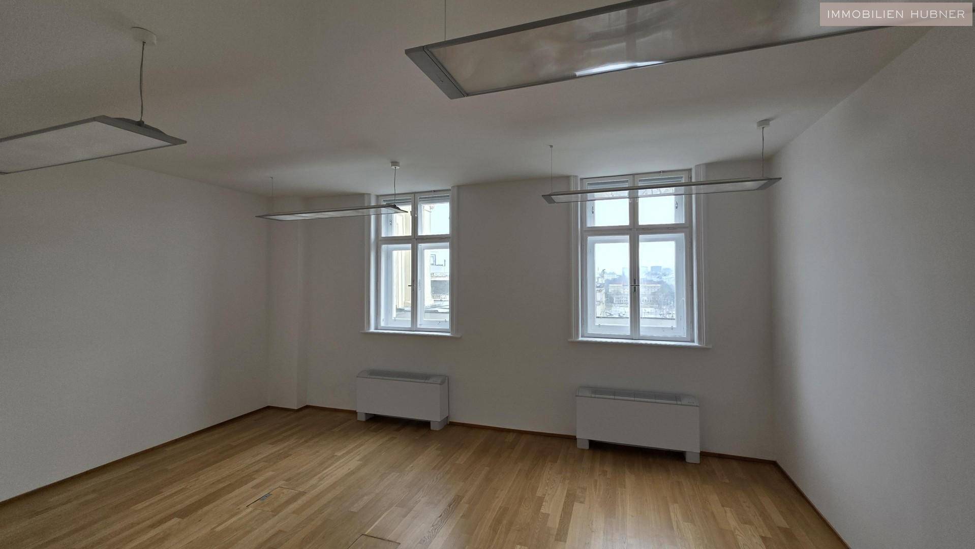 Helles, leeres Büro mit zwei großen Fenstern, Holzparkettboden und modernen Deckenleuchten.