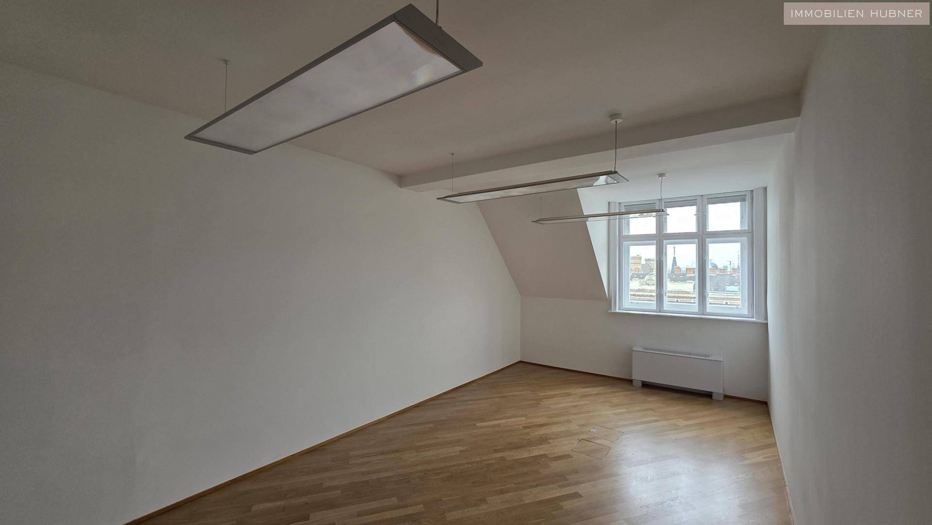 Leeres Büro mit Dachschräge, Holzparkettboden und einem Fenster, das viel Tageslicht hereinlässt.