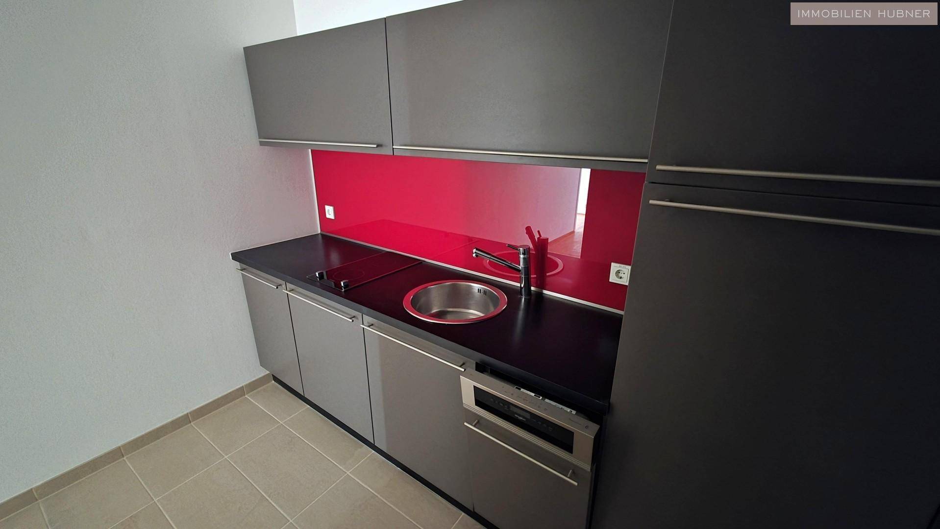 Kompakte Küchenzeile mit grauen Schränken, schwarzer Arbeitsplatte und einer roten Spritzschutzwand.