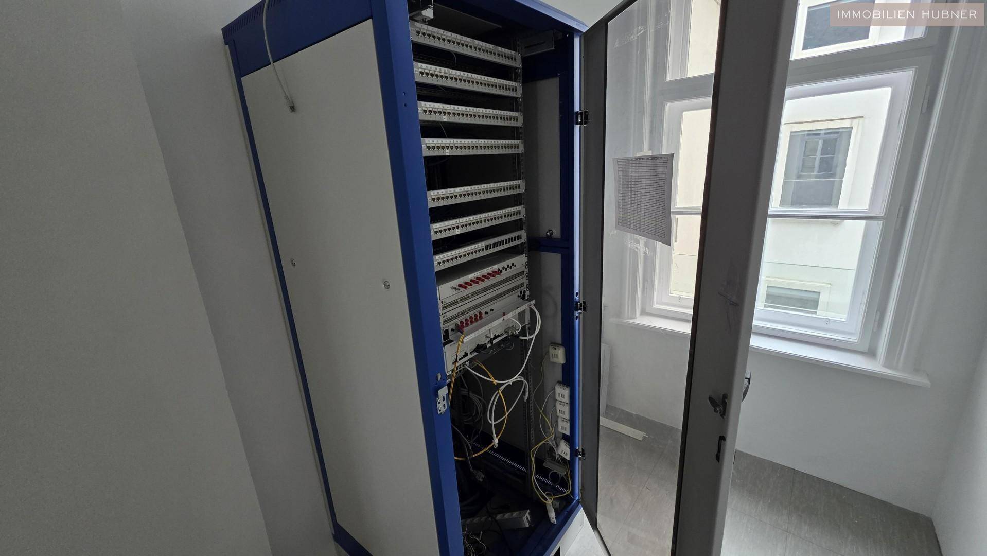 Offener Serverschrank mit Verkabelung und Netzwerkgeräten in einem Technikraum neben einem Fenster.