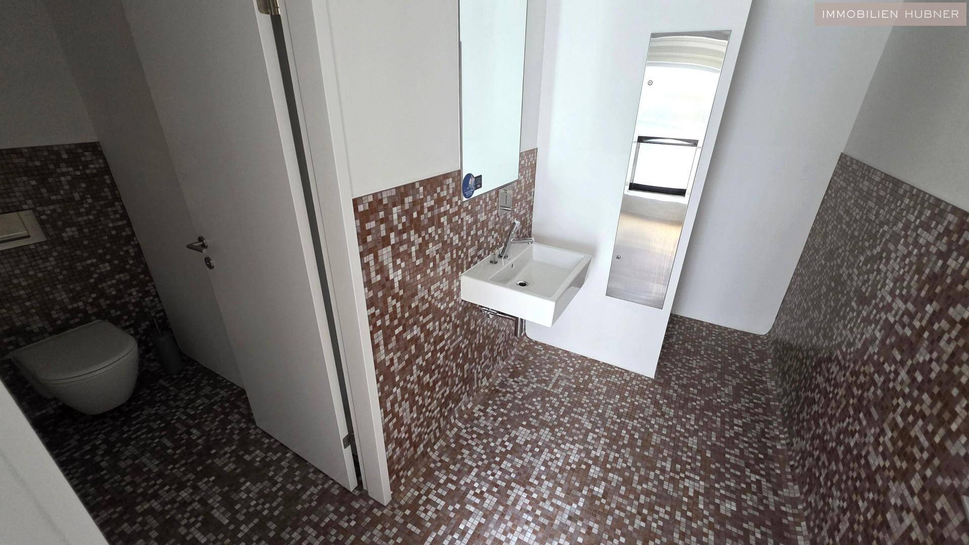 Modernes WC mit Mosaikfliesen, wandmontierter Toilette, Waschbecken und einem großen Spiegel.