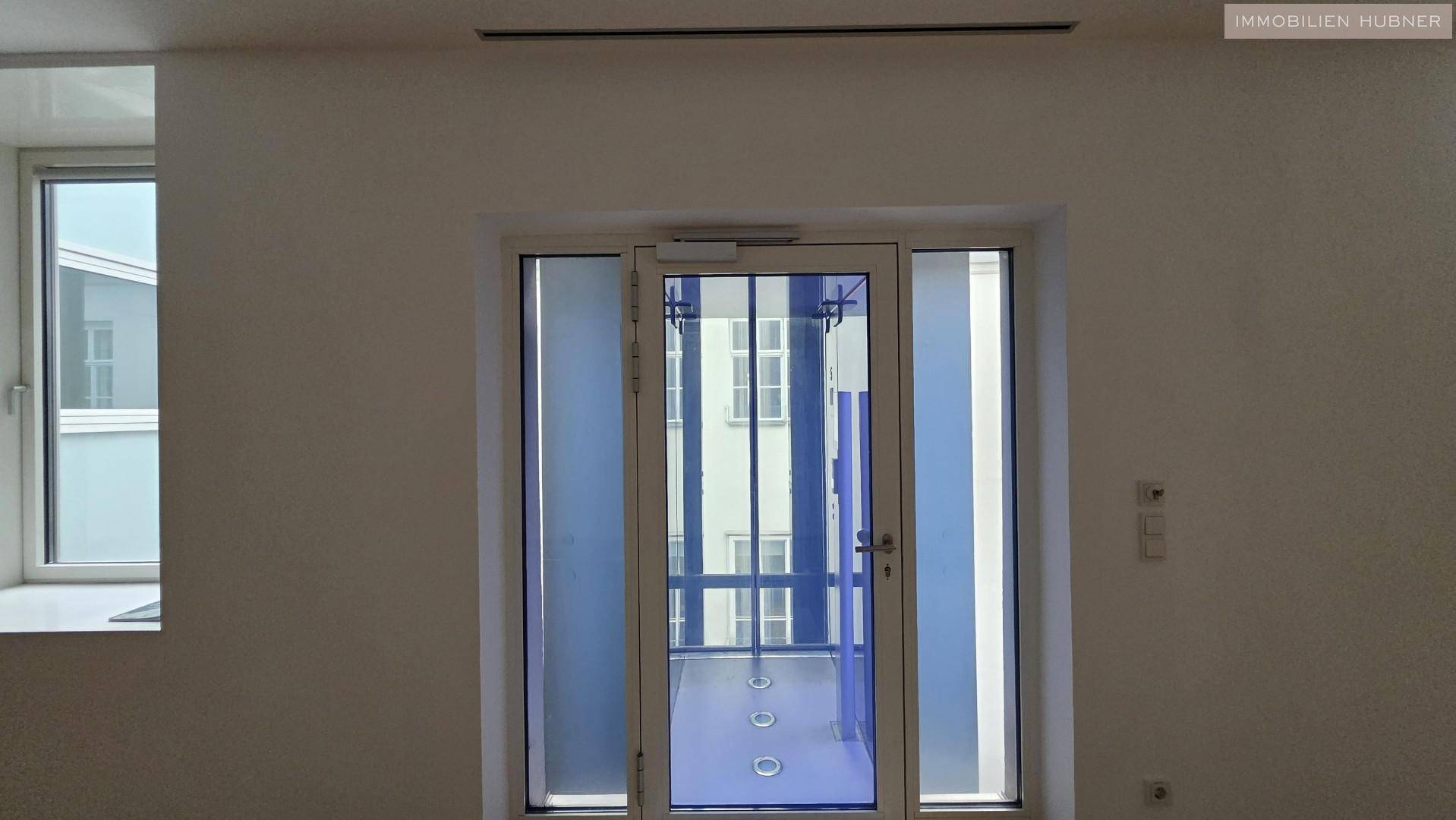 Innenansicht eines Flurs mit einem Fenster und einer modernen Aufzugstür mit blauer Verglasung.