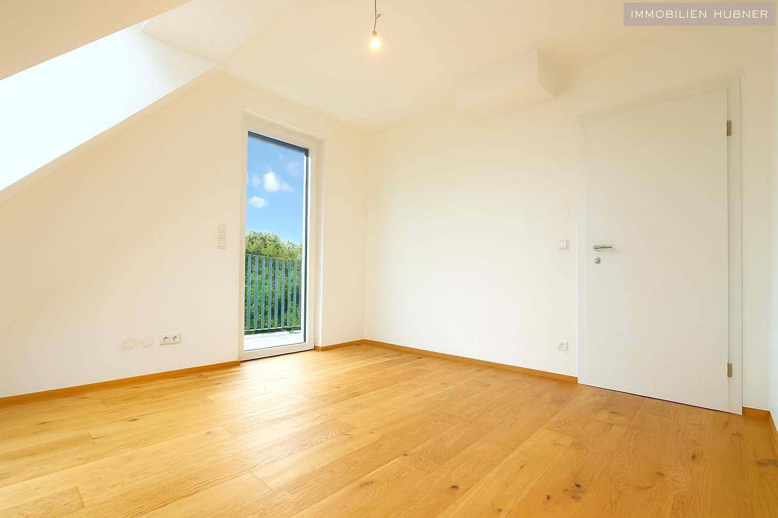 Freundliches Zimmer mit Holzboden, Dachschräge und Zugang zu einem kleinen Balkon mit Blick ins Grüne.