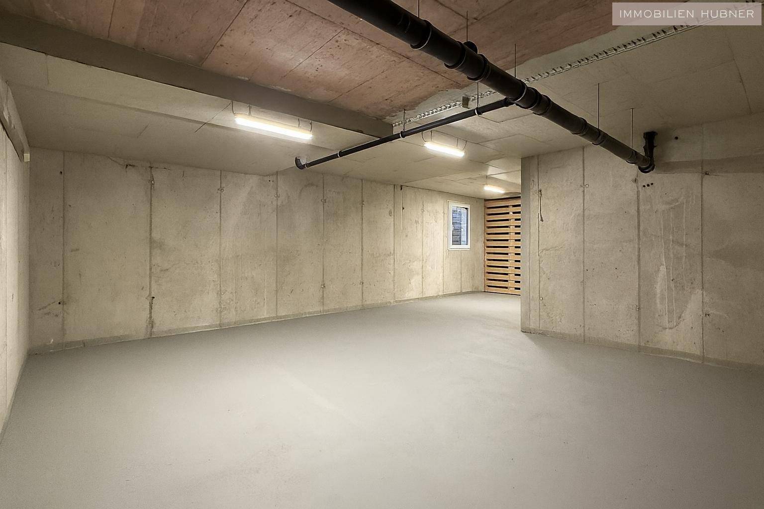 Kellerraum mit Betonwänden, Rohren an der Decke und einem kleinen Fenster.