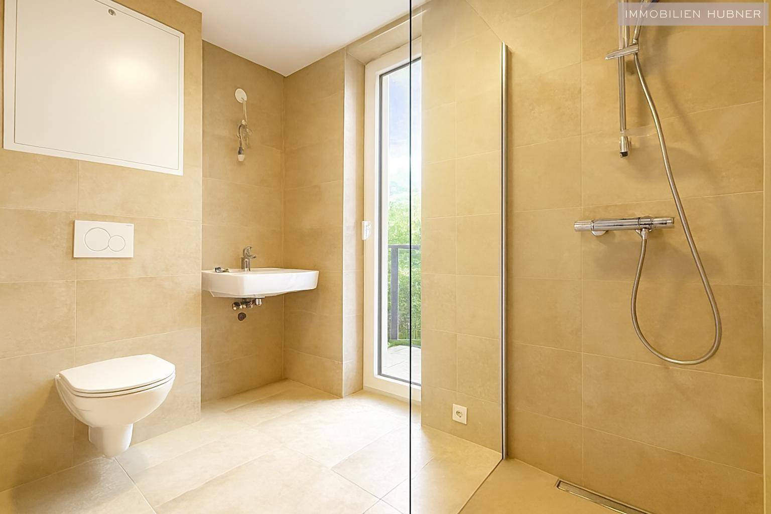 Modernes Badezimmer mit beigefarbenen Fliesen, wandhängendem WC und ebenerdiger Dusche mit Glaswand.