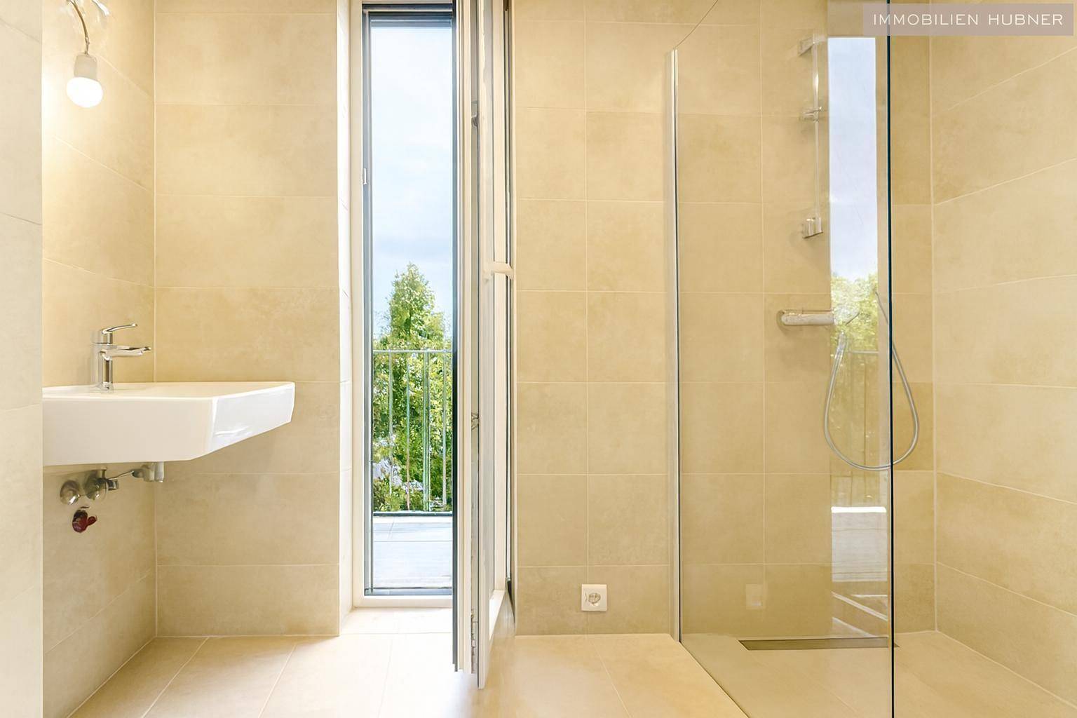 Helles Badezimmer mit beigefarbenen Fliesen, Waschbecken, ebenerdiger Dusche und Zugang zu einem Balkon.