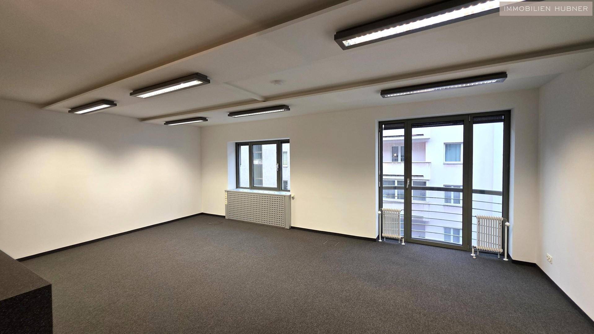 Heller Büroraum mit Teppichboden, großen Fenstern und Zugang zu einem möglichen Balkon oder einer Loggia.