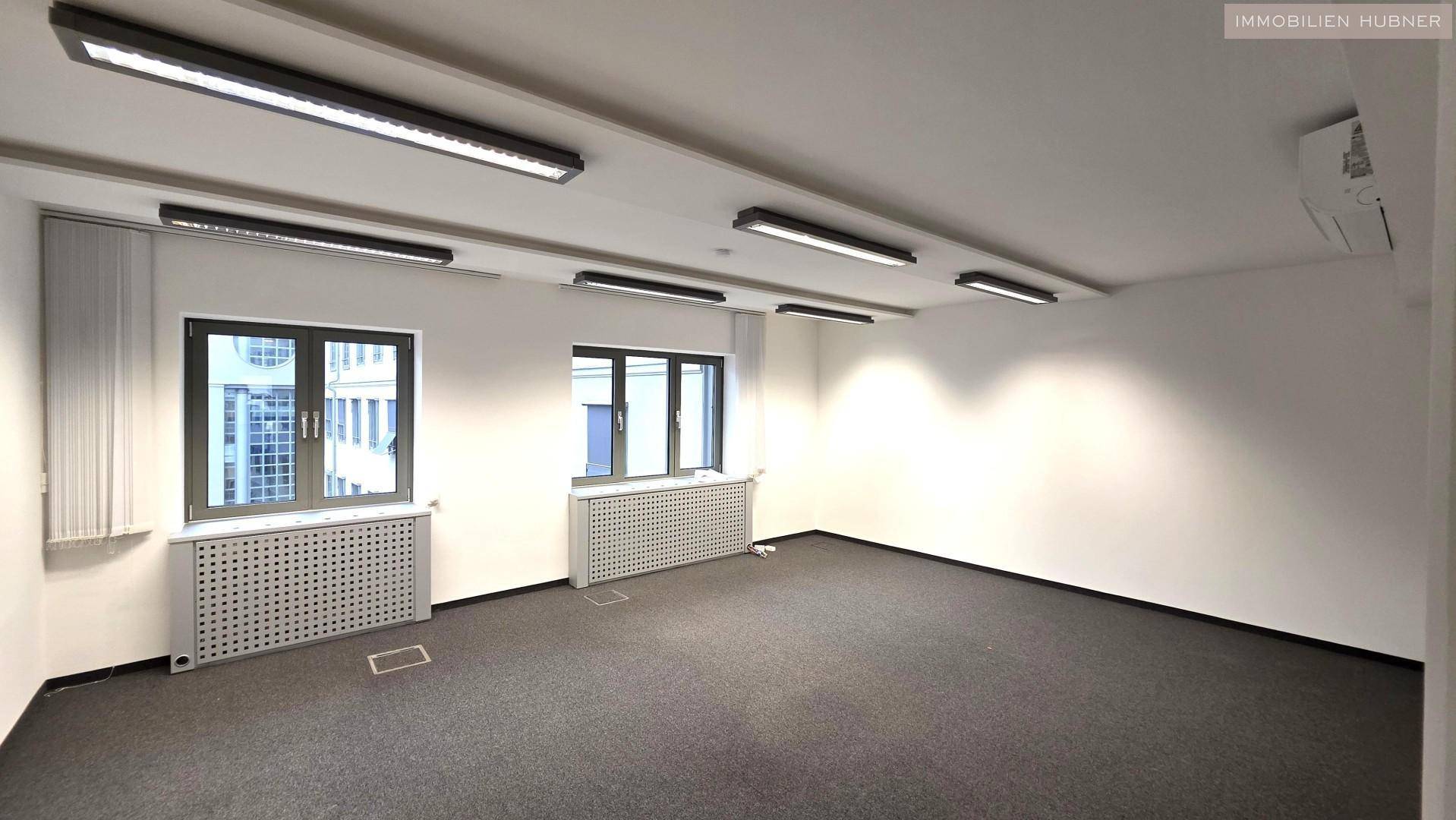 Heller Büroraum mit großen Fenstern, Teppichboden und effizienter Deckenbeleuchtung.