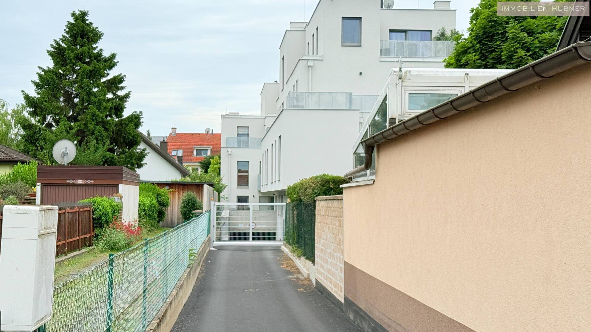 Zufahrt zur modernen Wohnanlage mit Zaun und angrenzenden Gärten, umgeben von Bäumen und Nachbarhäusern.