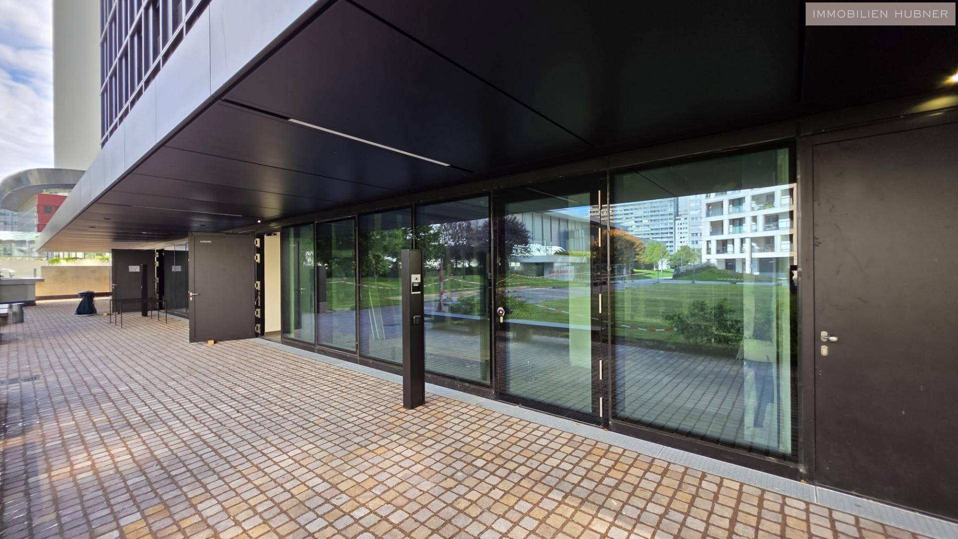 Eingangsbereich des modernen Gebäudes mit großen Glasfronten und gepflastertem Vorplatz.