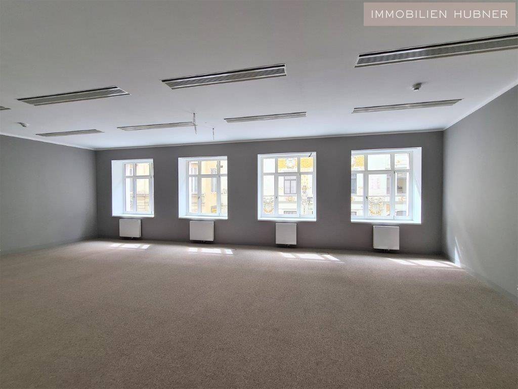Geräumiger, leerer Büroraum mit Teppichboden und mehreren Fenstern, die viel Tageslicht hereinlassen.