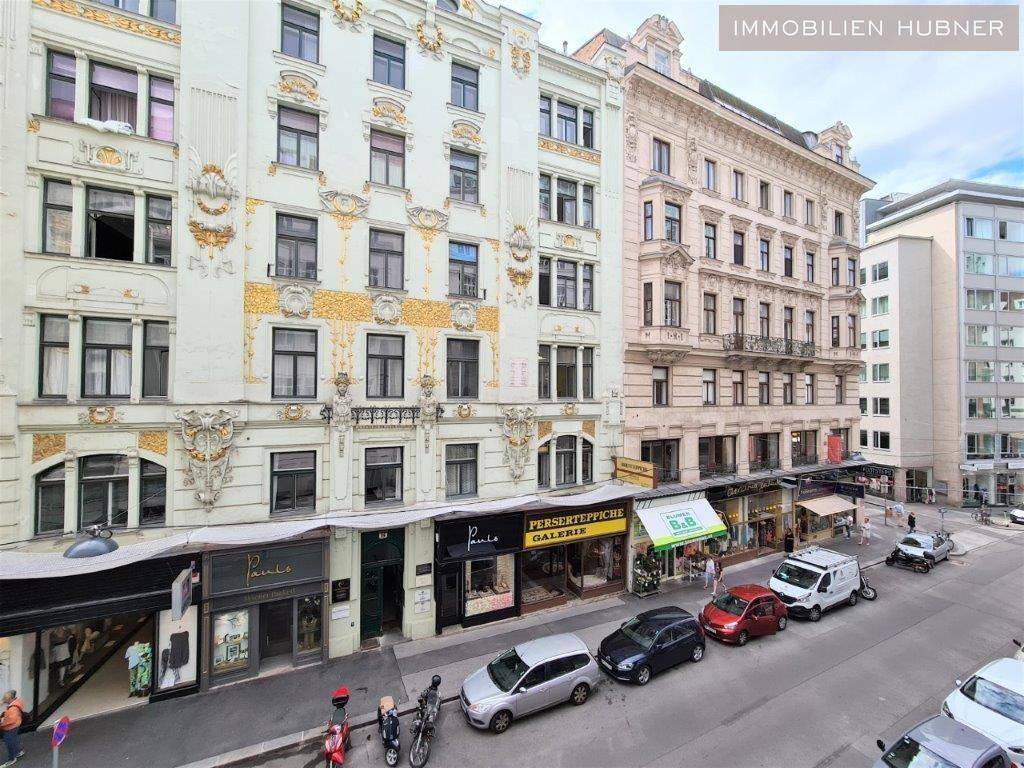 Historische Gebäude mit reich verzierten Fassaden entlang einer belebten Straße mit Geschäften und parkenden Fahrzeugen.
