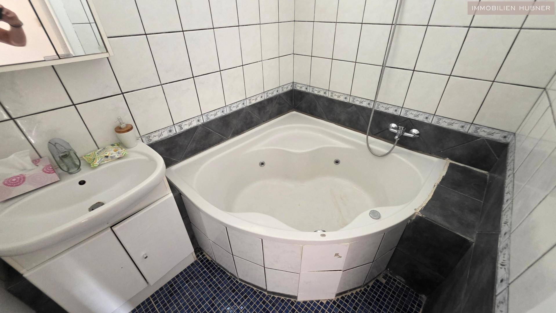 Badezimmer mit Eckbadewanne und Waschbecken, gefliest in Weiß und Dunkelgrau.