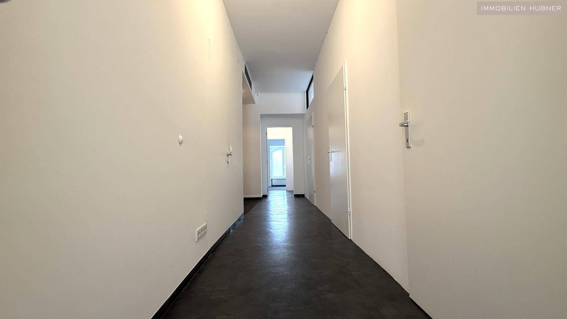 Schmaler, langer Flur mit dunklem Fliesenboden und weißen Wänden, der verschiedene Bereiche verbindet.