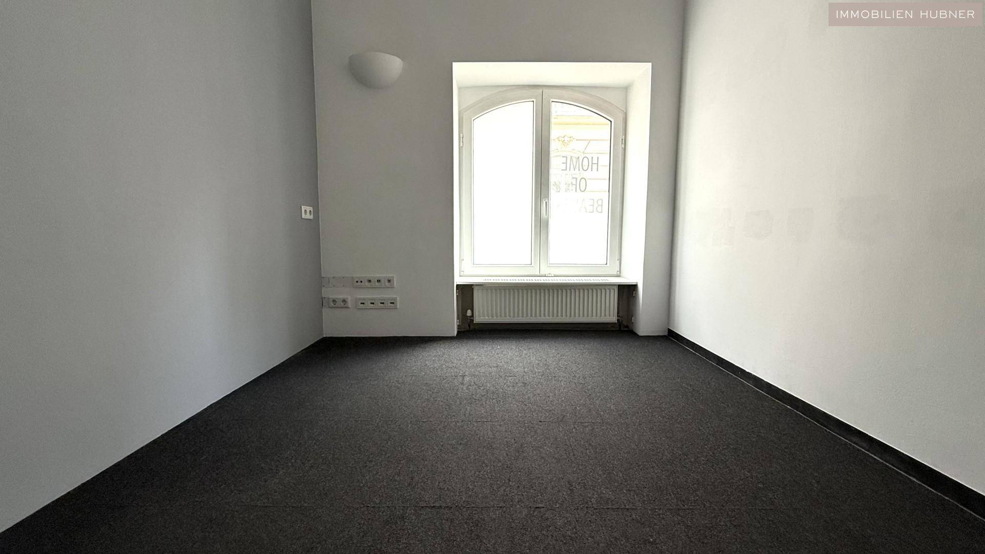 Schlichter Raum mit einem großen Bogenfenster, Teppichboden und weißen Wänden, bietet vielseitige Nutzungsmöglichkeiten.
