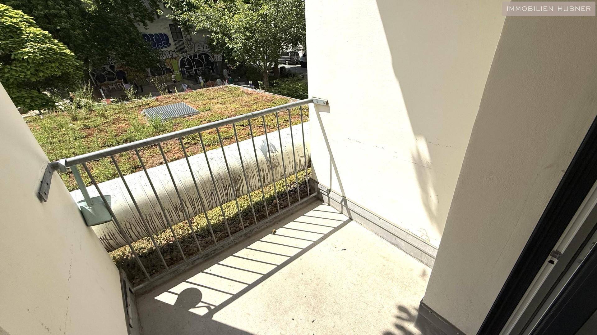 Kleiner Balkon mit Metallgeländer und Betonboden, bietet einen grünen Ausblick auf Dächer und Bäume.