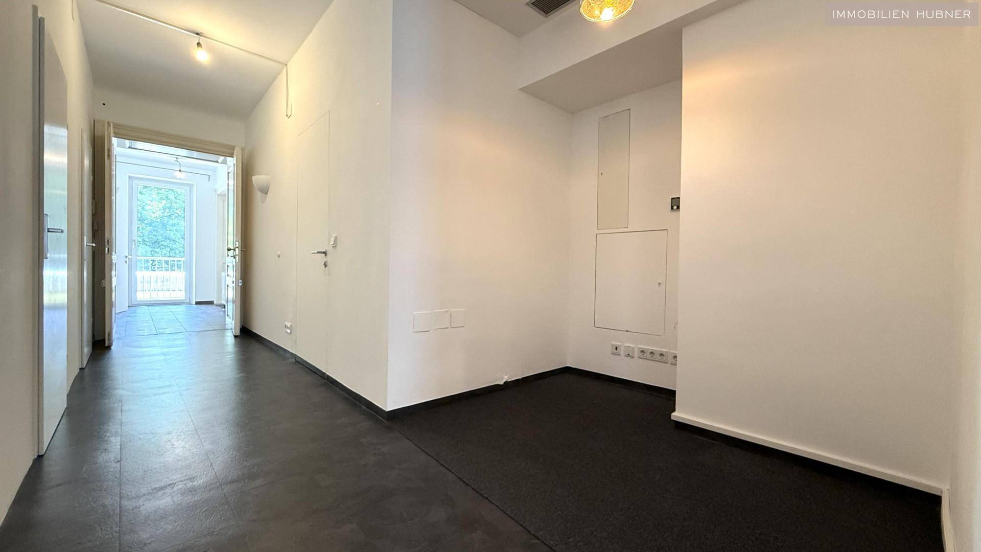 Langer Flur mit dunklem Fliesenboden und weißen Wänden, führt zu einem hellen Bereich mit Balkonzugang.