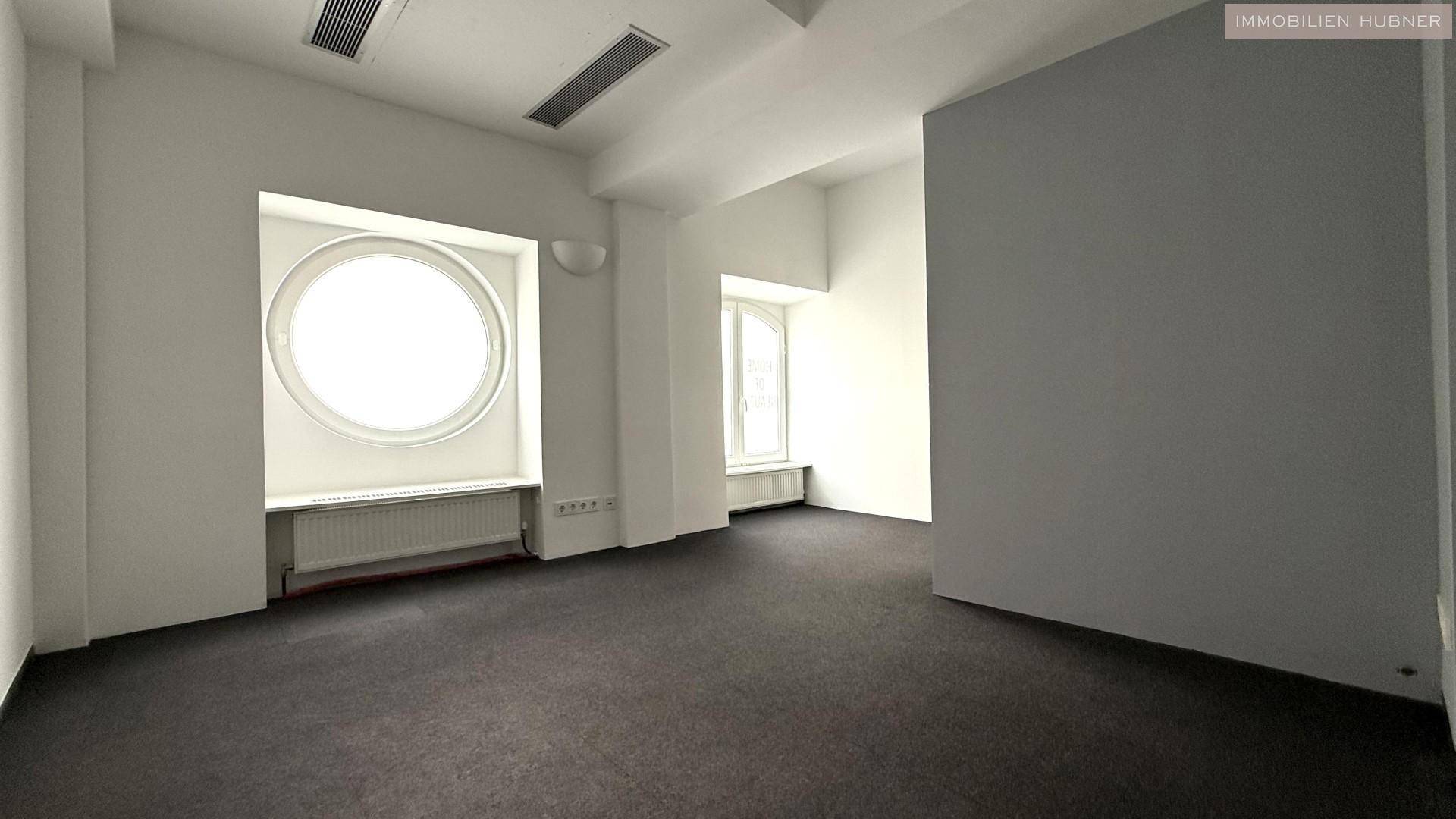 Heller Raum mit einem markanten runden Fenster und einem Bogenfenster, Teppichboden und Deckenlüftung.