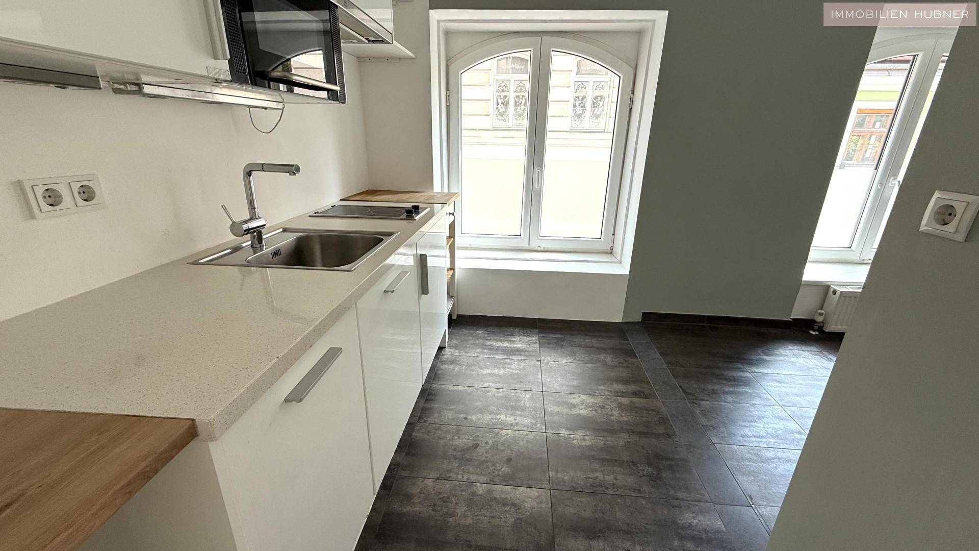 Moderne Küchenzeile mit weißen Schränken, Spüle und Mikrowelle, ergänzt durch ein Fenster und dunkle Fliesen.