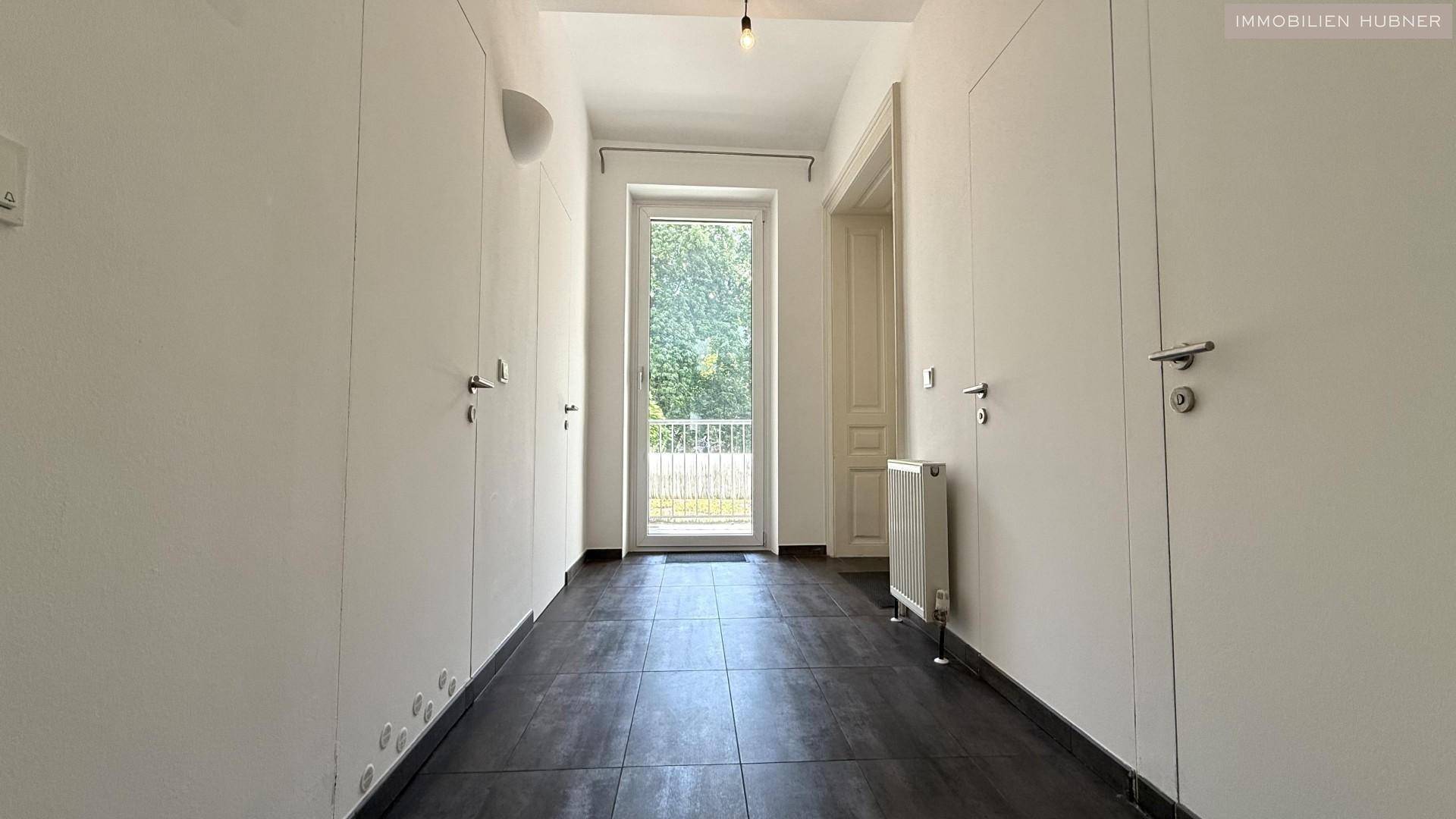 Heller Flur mit dunklem Fliesenboden und weißen Wänden, bietet direkten Zugang zu einem Balkon.
