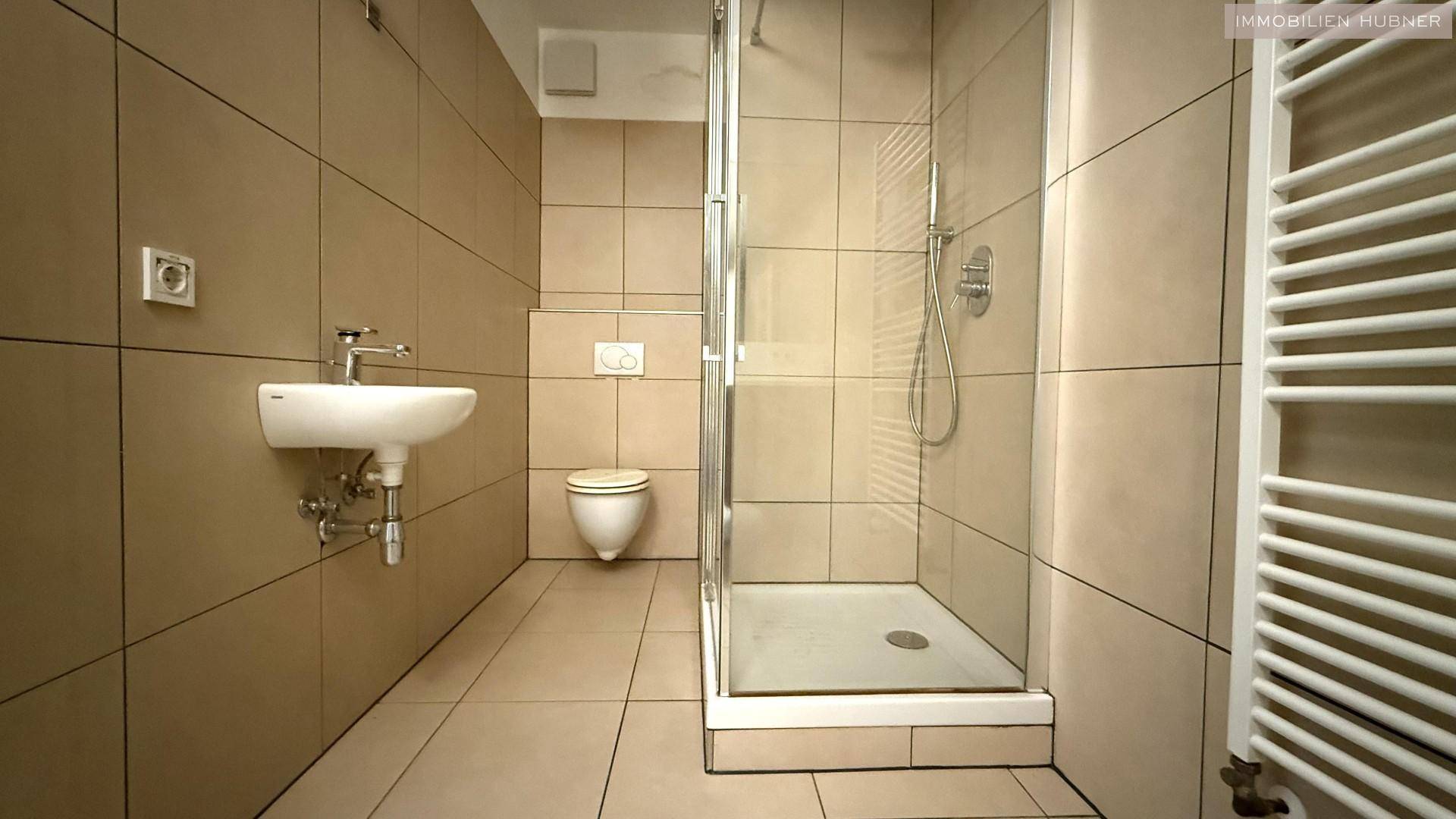 Badezimmer mit beigefarbenen Fliesen, einer Duschkabine, Waschbecken und wandhängender Toilette sowie Handtuchheizkörper.