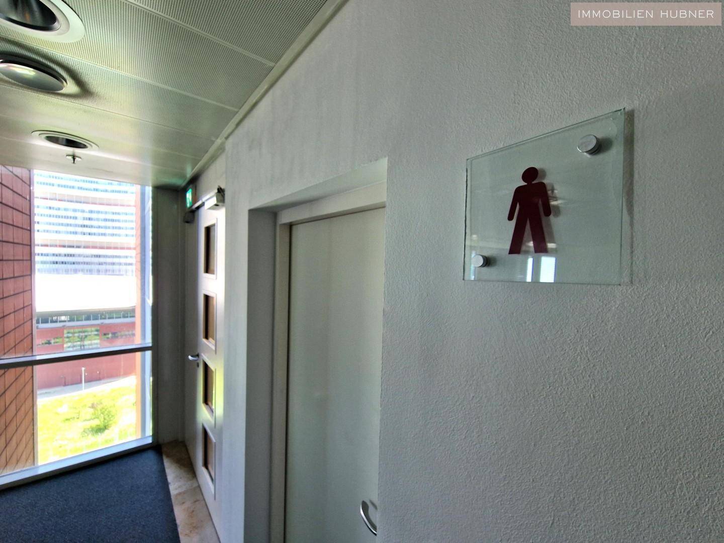 Flur mit Zugang zu den Toiletten, gekennzeichnet durch ein Piktogramm, und Fenster mit Ausblick.