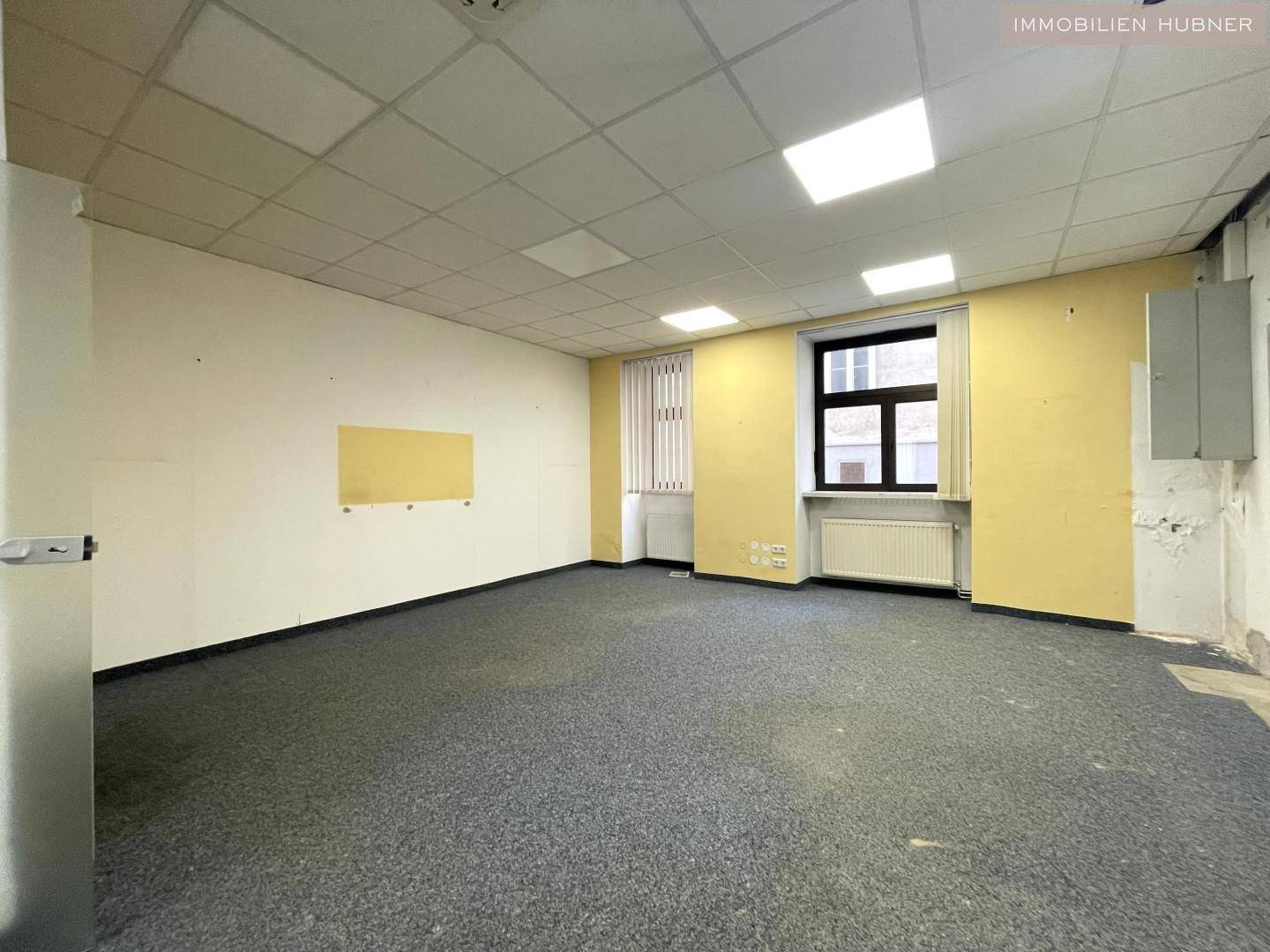 Leeres Bürozimmer mit Teppichboden, Rasterdecke und einem Fenster mit Heizkörper, teilweise renovierungsbedürftig.