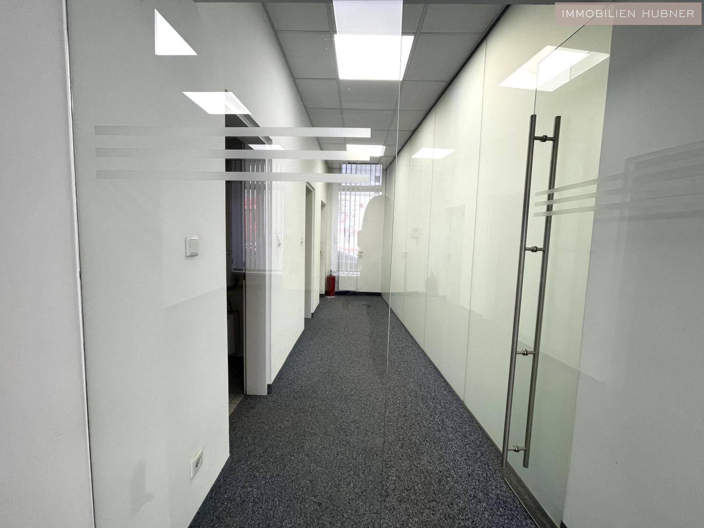 Moderner Büroflur mit Glaswänden, Teppichboden und einer Rasterdecke, der zu weiteren Räumen führt.