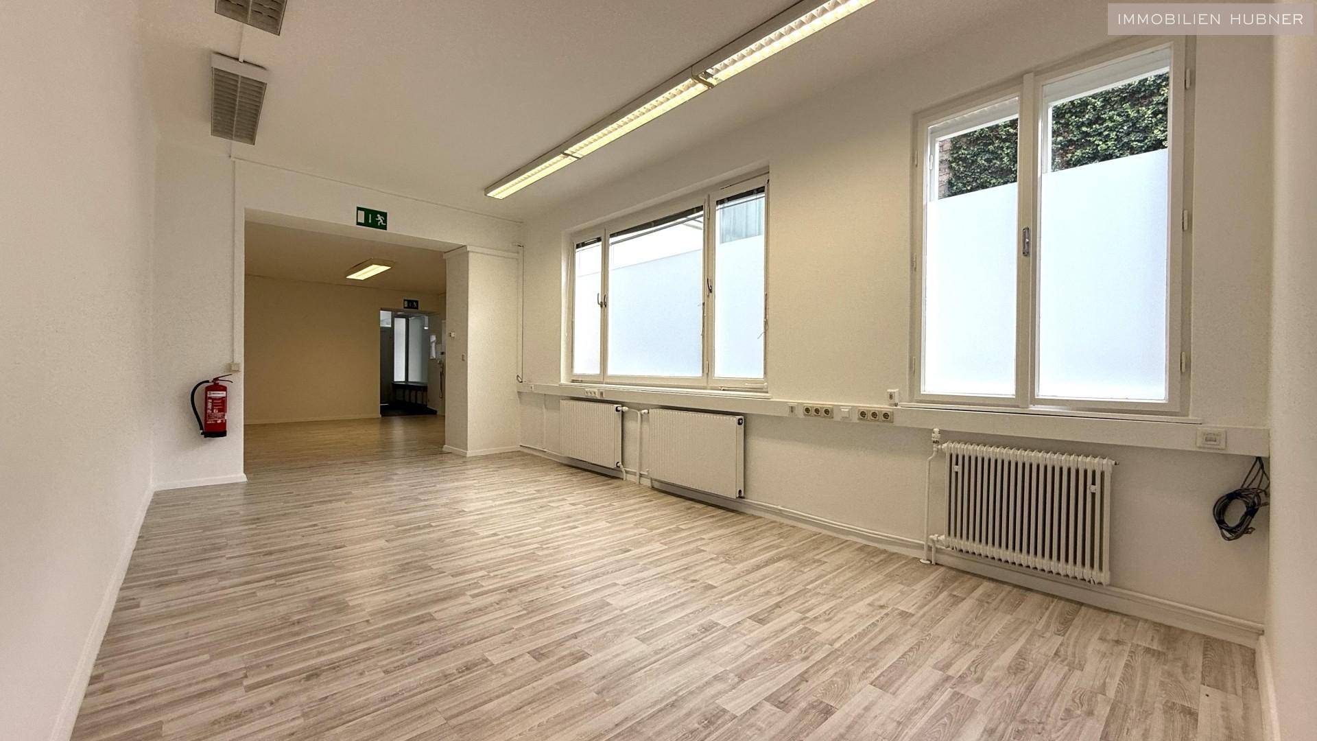 Geräumiger Büroraum mit hellem Laminatboden, großen Fenstern und Heizkörpern unter den Fenstern.