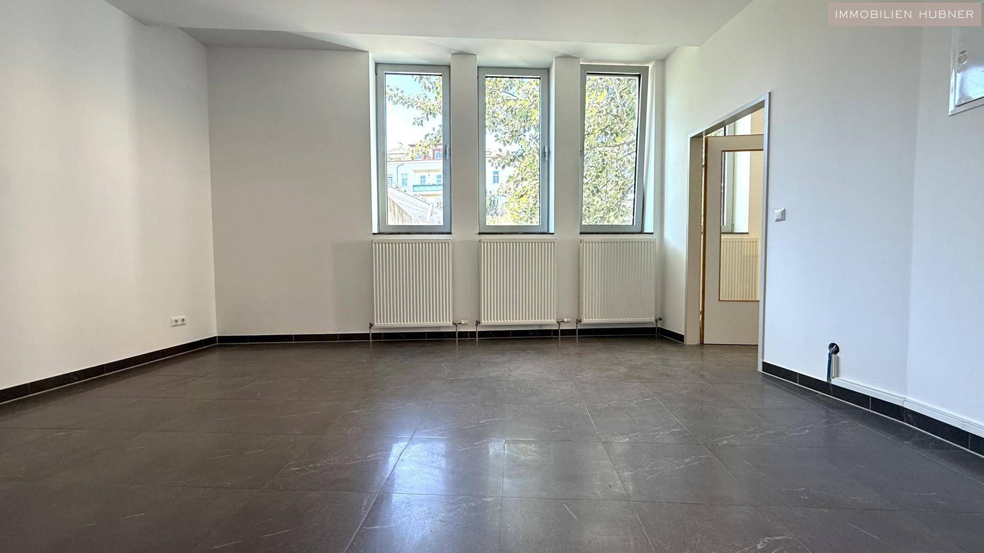 Heller Büroraum mit Fliesenboden, Heizkörpern unter den Fenstern und einer Tür.