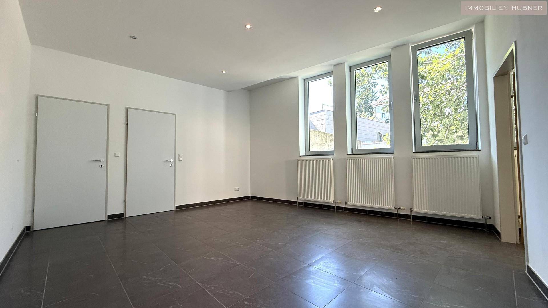 Heller Büroraum mit drei hohen Fenstern, Fliesenboden und weißen Türen.